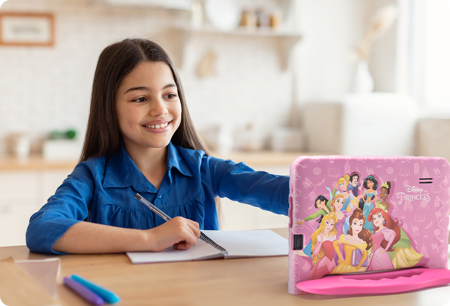 Foto de uma menina sentada em uma cadeira com uma caneta e um caderno, e à sua frente, o Tablet Princesas.